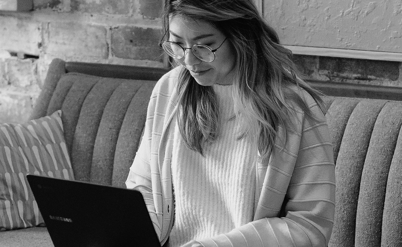 A black and white photo of an Asian woman with long hair wearing glasses and a light sweater under a cardigan is typing on her laptop while smiling.